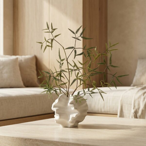 Dans un salon beige apaisant, le vase minimaliste ornemental en céramique blanc aux formes ondulées repose sur une table, avec fines branches vertes décoratives.