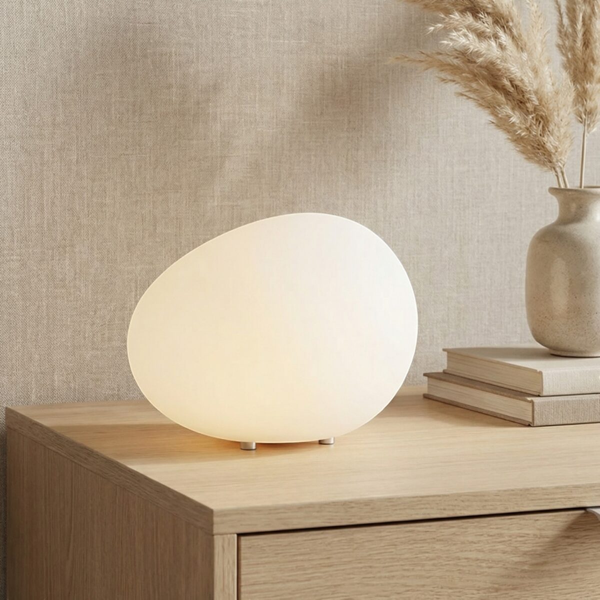 La lampe à poser Japandi de forme organique blanche posée sur meuble bois clair, galet lumineux LED diffusant lumière douce, ambiance zen minimaliste chaleureuse.