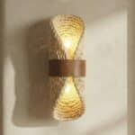 Applique murale japandi rotin tressé beige à la forme verticale, cerclage en cuir taupe avec deux ampoules LED allumée sur un mur enduit chaud.