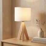 La lampe japandi bois clair et abat-jour beige allumée sur commode chêne clair avec vase céramique beige et brindilles séchées.
