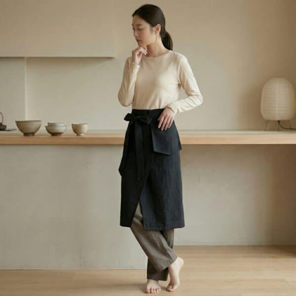 Une jeune femme portant un tablier japonais de taille à nouer de couleur noire sur un haut beige et un pantalon fluide, dans une cuisine lumineuse et épurée au style minimaliste.