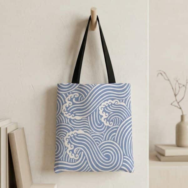 Accroché à un crochet mural, le Sac cabas japonais motif vagues bleu montre un grand motif de vagues blanches sur fond bleu avec anses noires dans un décor intérieur minimaliste.