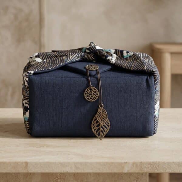 Sur une table en pierre claire, le sac japonais matelassé service thé vagues Hokusai bleu est noué avec un tissu à motif de vagues et décoré d’un pendentif feuille en métal.