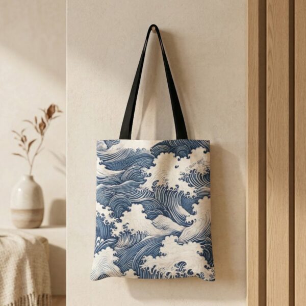 Accroché à un mur clair, le sac japonais en toile grande vague Hokusai montre un motif d’estampe bleue représentant de grandes vagues avec anses noires dans un intérieur minimaliste.