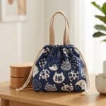 Sur une table en bois, le sac lunch japonais en coton motif chat bleu à cordon montre des dessins de chats blancs. Petit sac bento décoratif posé près d’une tasse et d’une plante.