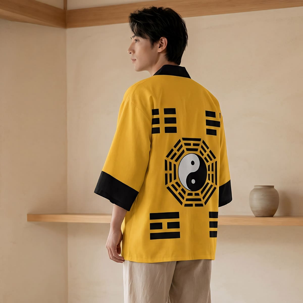 Un homme de dos portant une veste haori homme jaune avec symbole yin et yang noir et blanc au centre de motifs géométriques, dans une pièce épurée au style japonais avec des murs clairs et du mobilier en bois clair.
