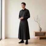 Un jeune homme portant un pantalon japonais homme coupe hakama noir avec une chemise noire, posant dans une pièce lumineuse au décor épuré avec un vase et des branches sèches.