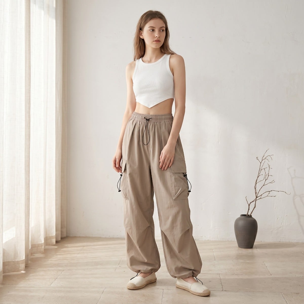 Une jeune femme caucasienne portant le pantalon parachute femme cargo avec cordons, beige, dans une pièce à vivre claire et ensoleillé.