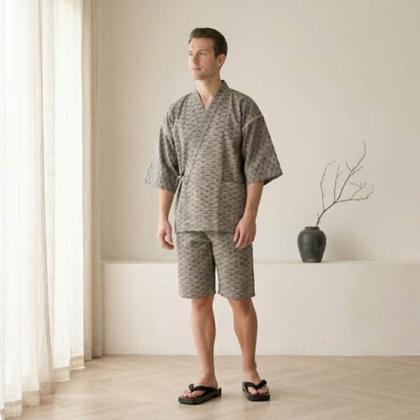 Jeune homme de type caucasien en jinbei homme motif seigaiha coton taupe, veste croisée à manches courtes et short assorti, debout dans un salon japandi clair.