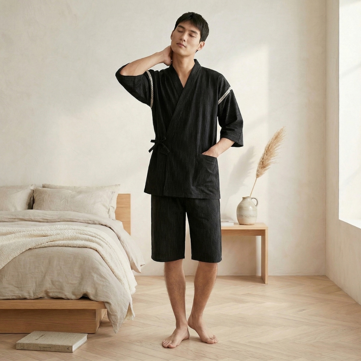 Un homme brun portant un pyjama japonais à fines rayures et avec short pour homme de couleur noire dans une chambre lumineuse et épurée avec un lit en bois clair.
