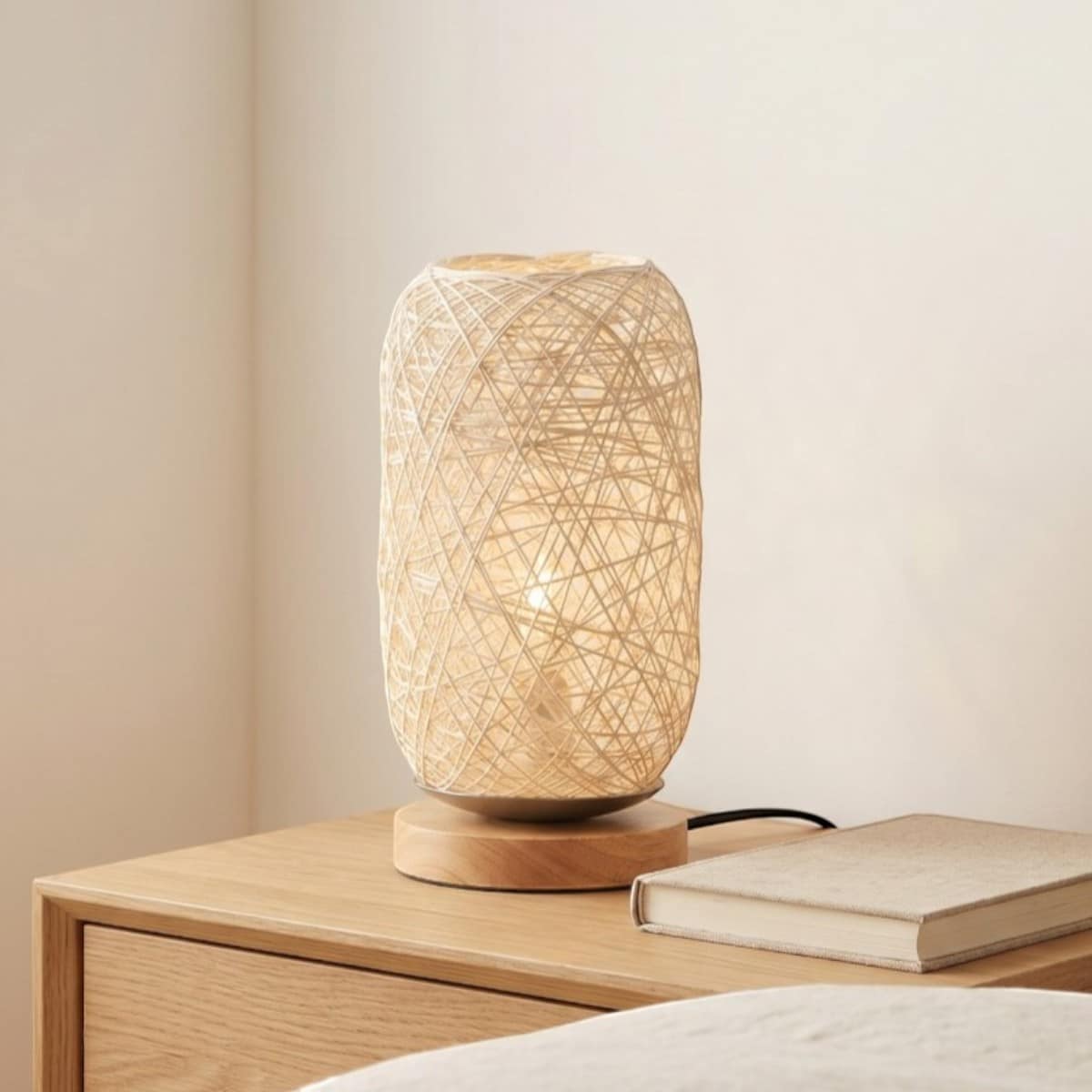 Lampe japandi corde chanvre tressée effet cocon lumière posée sur socle bois rond, sur table de chevet en bois clair.