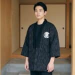 Un homme portant un Hanten homme avec symbole de prospérité noir à fines rayures, devant l’entrée d’une maison japonaise en bois.