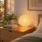 Une lampe japandi de table habillée de papier de riz, allumée sur une table de chevet en bois, avec des livres, une plante et un lit en arrière-plan.