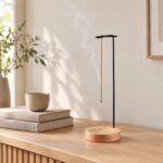 Un porte-encens japonais à potence noire sur socle rond, posé sur un meuble en bois avec un bâton d’encens fumant, près de livres et d’une plante.