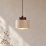 Une suspension Japandi en travertin large au bois foncé, avec un abat-jour cylindrique en pierre claire et un socle en bois brun, éclairant un mur beige près d’une branche décorative.